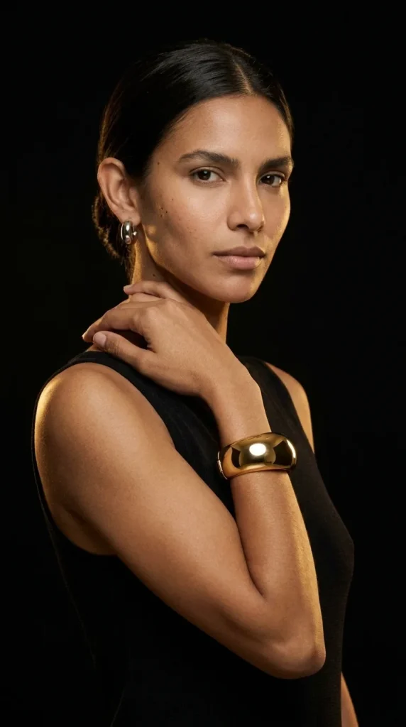 Mujer de negro con brazalete rígido dorado en la muñeca sobre fondo oscuro